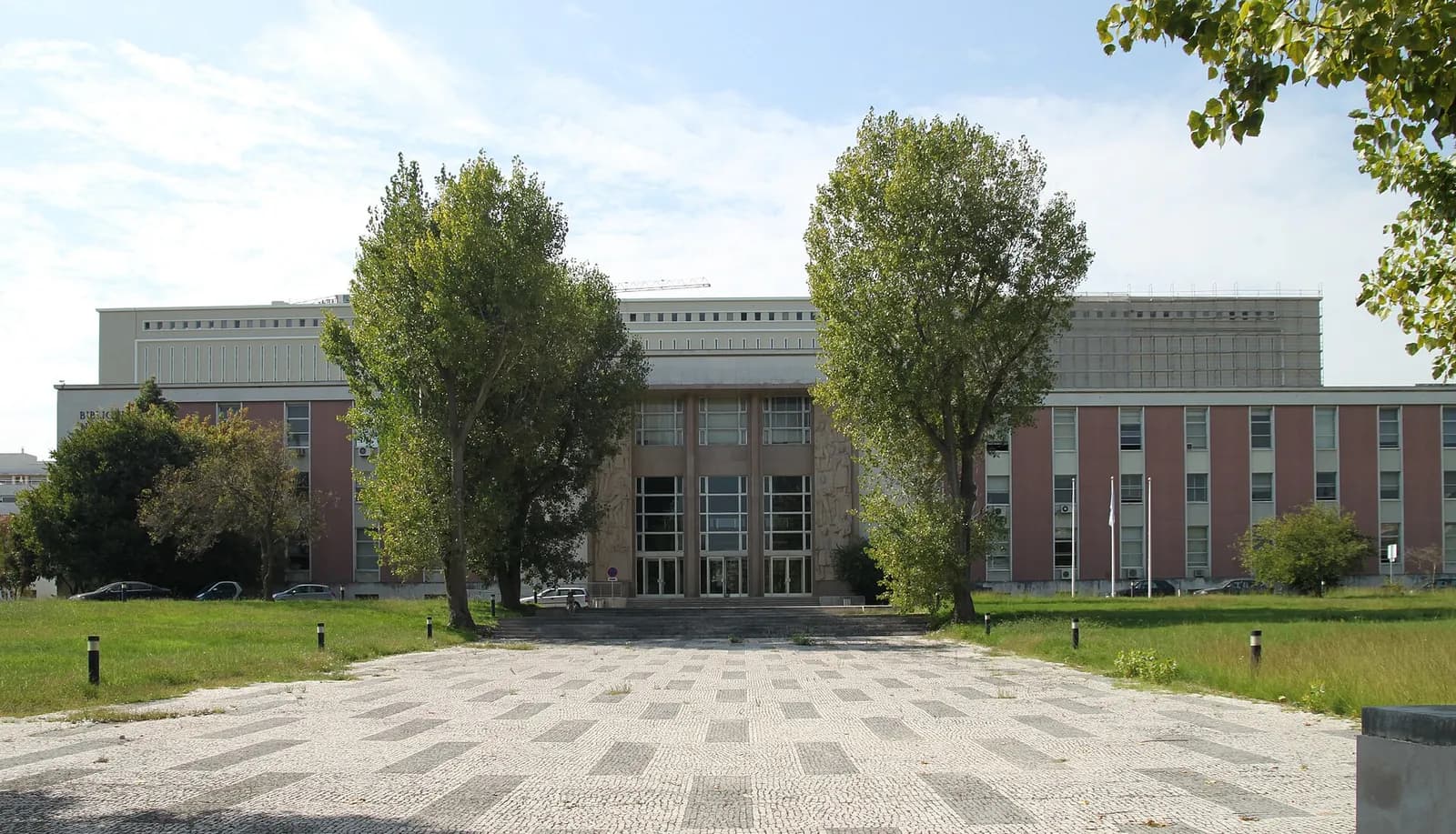 A long, low modernist building with pink-red panels between rows of windows and a taller pale block behind it, seen across a wide patterned stone forecourt. Two tall trees frame the glazed entrance, which is flanked by carved stone reliefs.