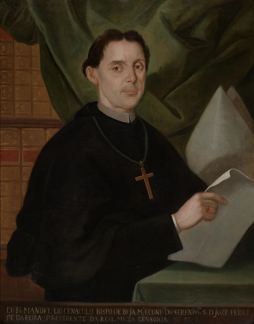Oil portrait of a dark-haired man in a black clerical habit with a pectoral cross, holding a sheet of paper beside a table. Shelves of leather-bound books fill the left background and a bishop's mitre stands on the right. An inscription runs along the bottom edge.
