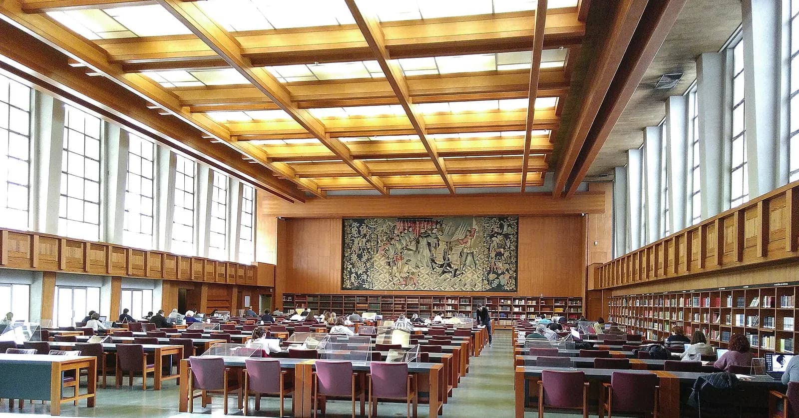 A long, high reading room with a coffered wooden ceiling lit from above and tall windows down both sides. Rows of desks with purple chairs, many occupied by readers, lead towards a large figurative woven hanging on the end wall above low bookshelves.