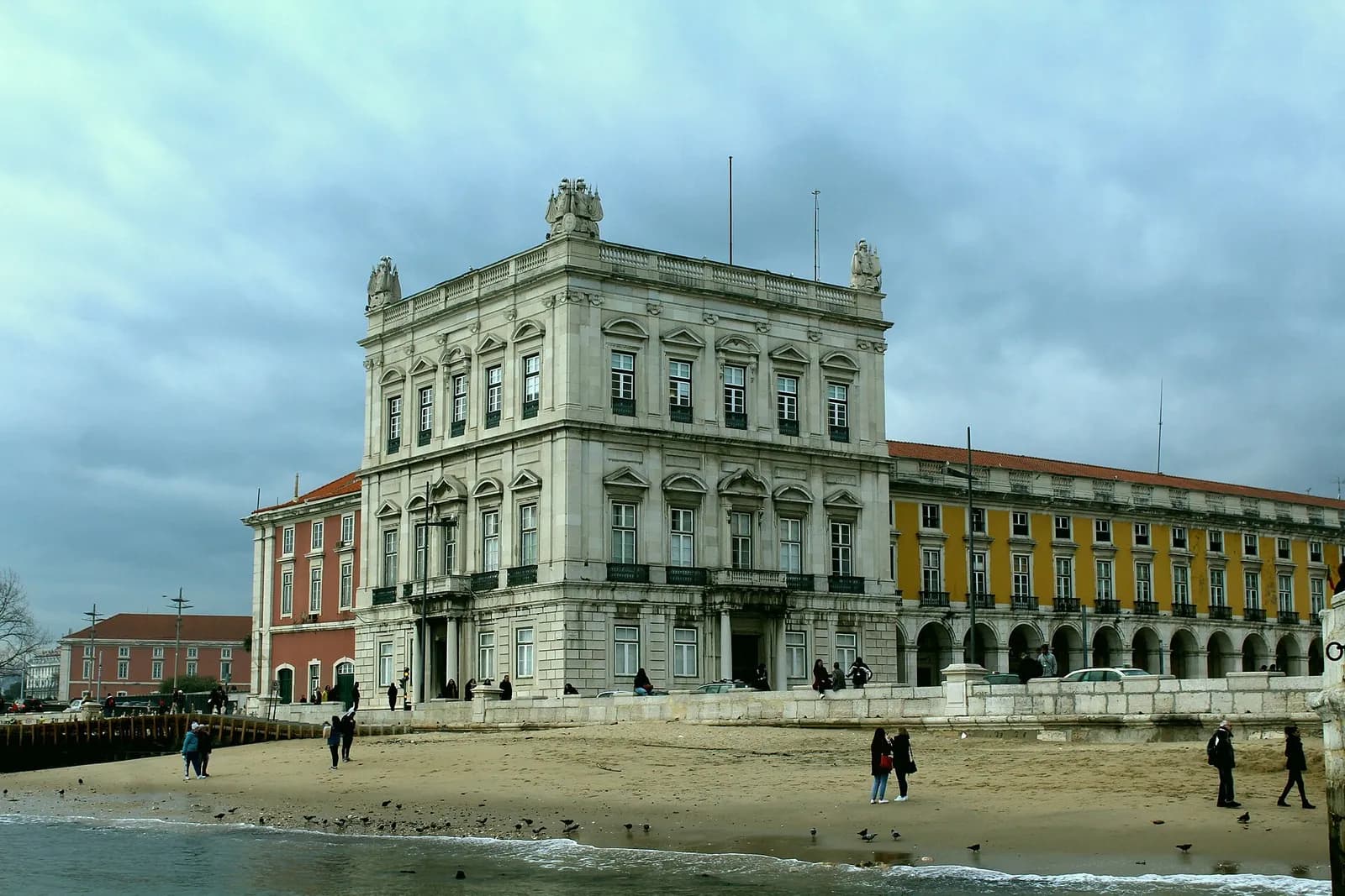 A square stone pavilion with balustrades and sculpted trophies on its roof stands at the corner of a long yellow arcaded building, behind a strip of sand at the river's edge where people walk under a cloudy sky.