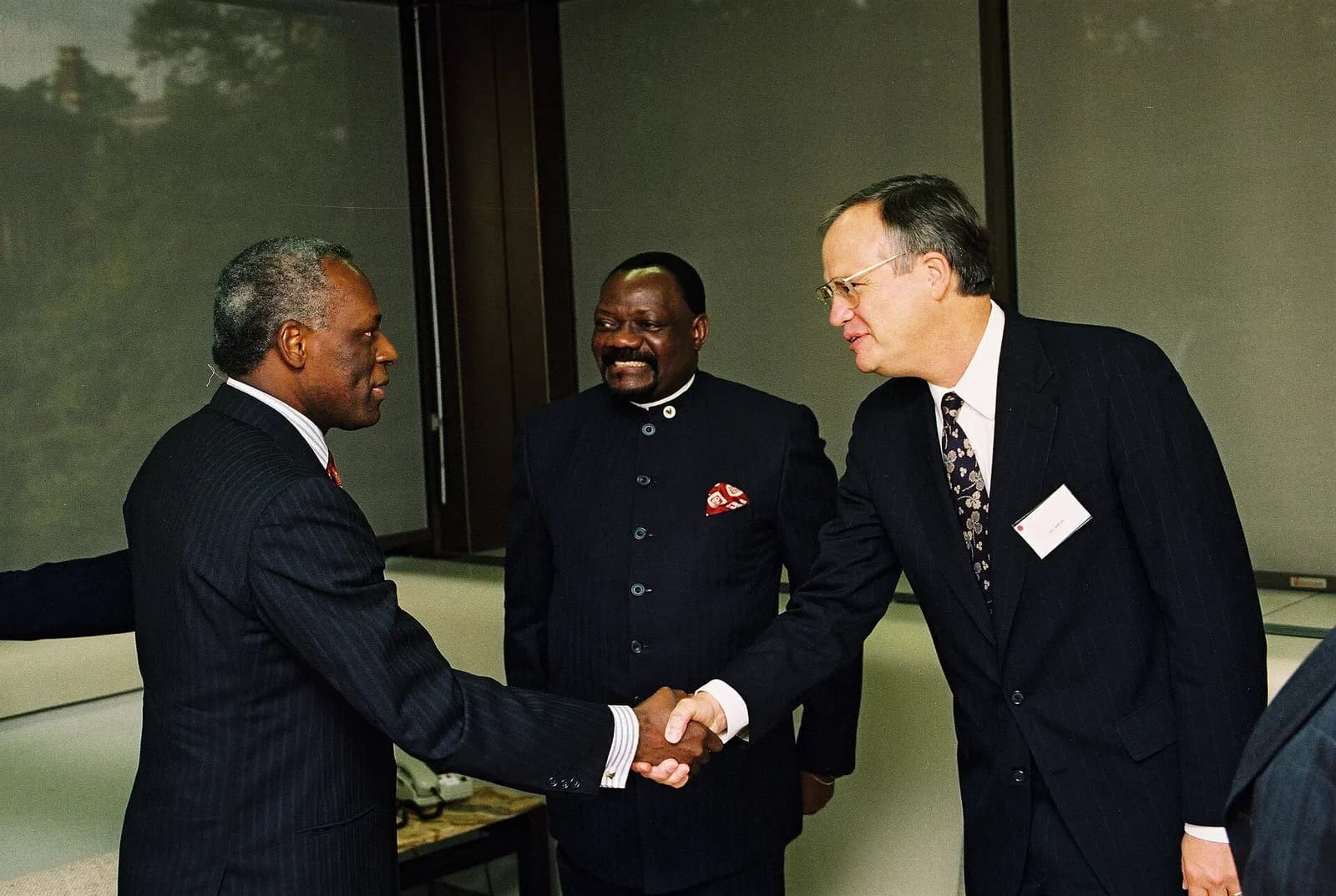 Three men in dark clothes in an office. On the left a grey-haired man shakes hands with a taller man in glasses wearing a name badge, while a smiling bearded man in a high-collared jacket with a red pocket handkerchief stands between them.