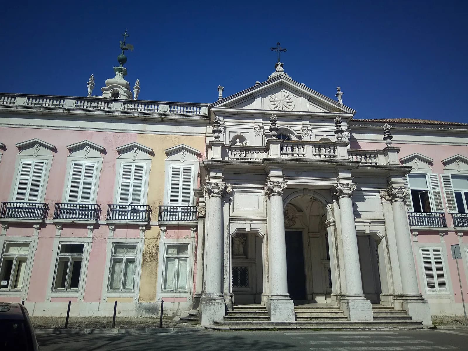 The front of a long two-storey palace with pink walls, white shuttered windows and iron balconies, and a white stone portico with columns and a pediment topped by a cross, under a deep blue sky.
