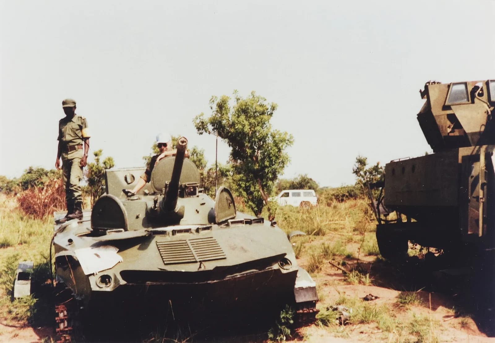 A tracked armoured vehicle with a small turret and gun sits in scrubland. An Angolan soldier in olive uniform stands on its hull and a man in a pale cap leans out of the hatch; a white vehicle marked UN is parked among the trees behind.