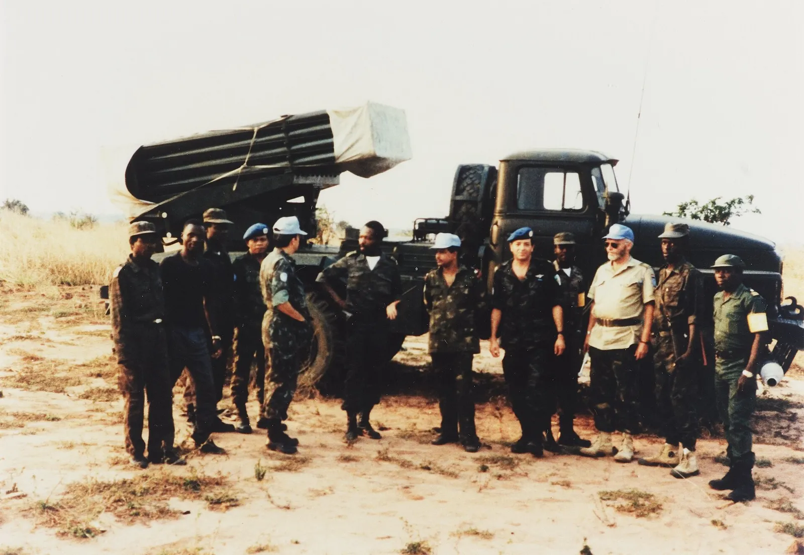 About a dozen men in uniform stand in a line on dry, sandy ground in front of a military truck carrying a multiple rocket launcher under a tarpaulin. Several wear UN blue berets or pale blue caps; the others wear Angolan camouflage or olive uniforms.