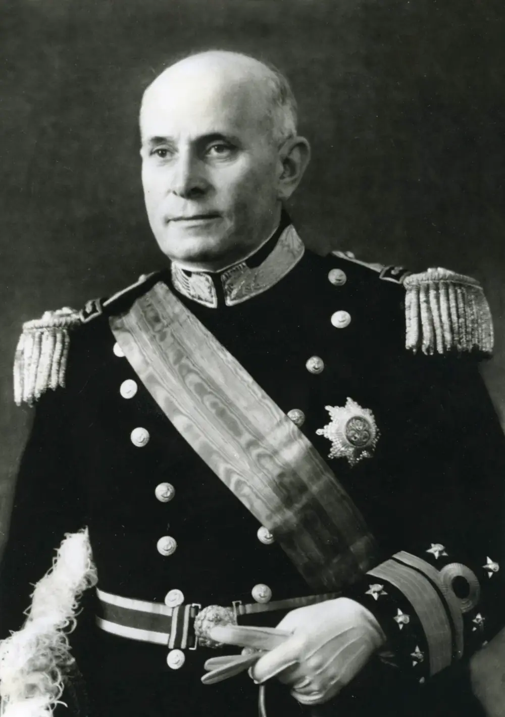 Black-and-white formal portrait of a bald older man in a dark naval dress uniform with fringed epaulettes, a broad sash across his chest and a star-shaped decoration, holding white gloves.