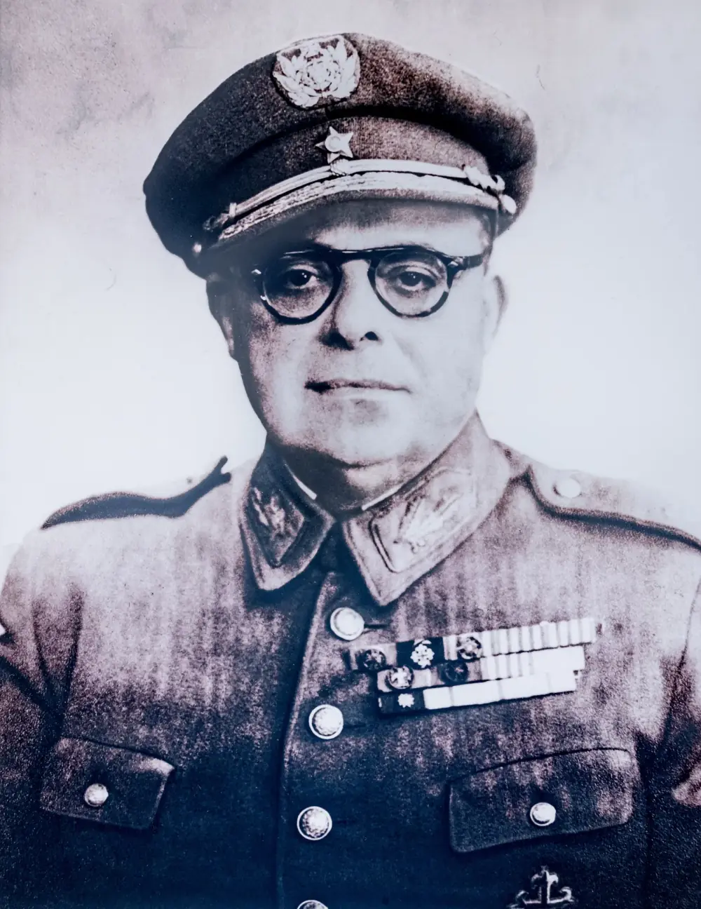Head-and-shoulders photograph of a heavy-set man in round dark-rimmed glasses, a peaked army cap with a large badge and a uniform jacket with rows of medal ribbons.