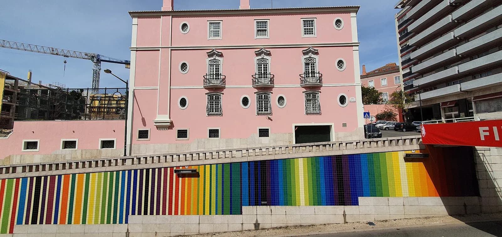 A three-storey pink palace with white window frames and small iron balconies stands above a long retaining wall tiled in vertical stripes of every colour. A crane and a modern apartment block flank it under a blue sky.