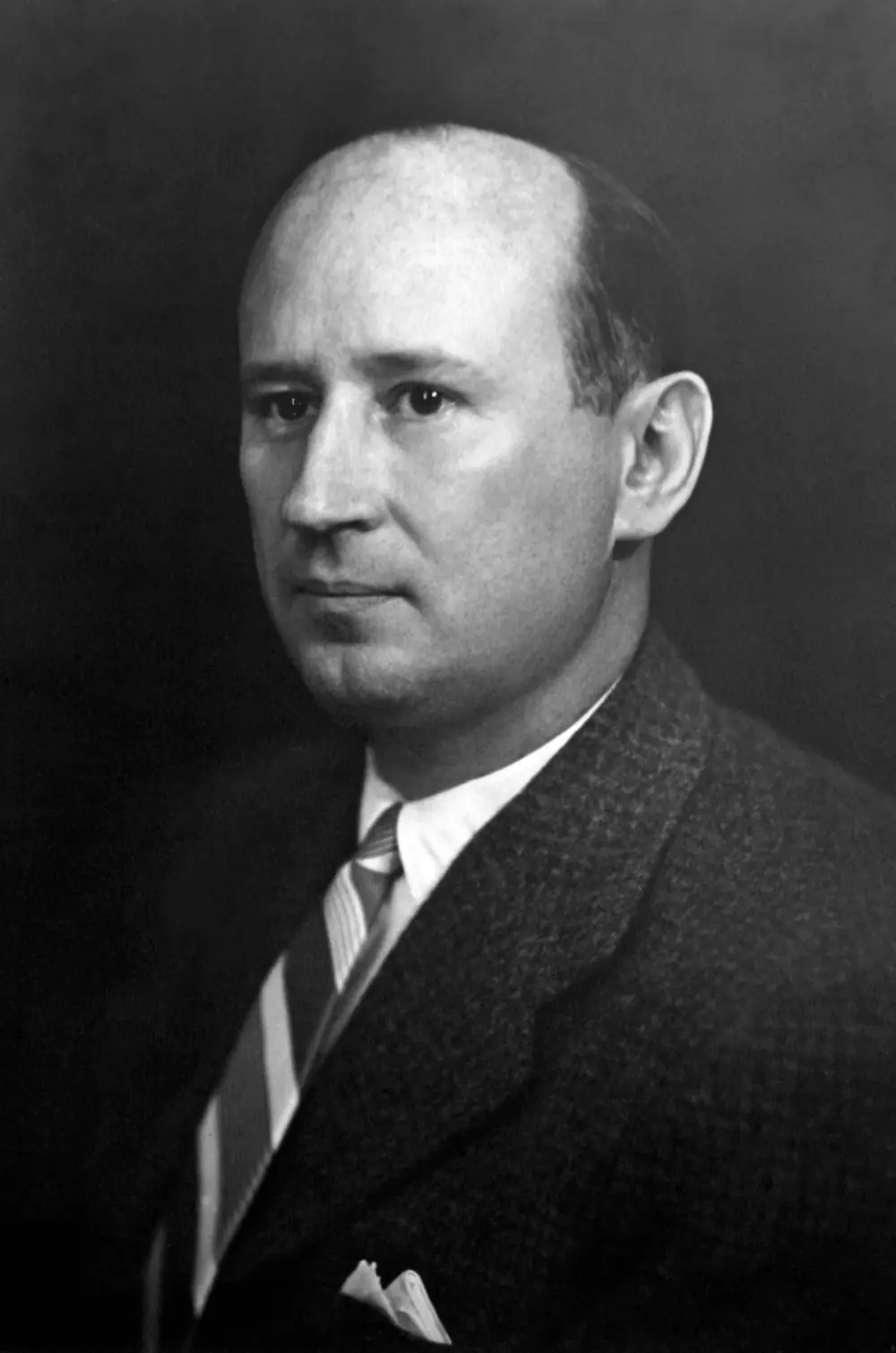Black-and-white studio portrait of a balding man in a dark tweed jacket, striped tie and pocket square, looking to the left of the camera.