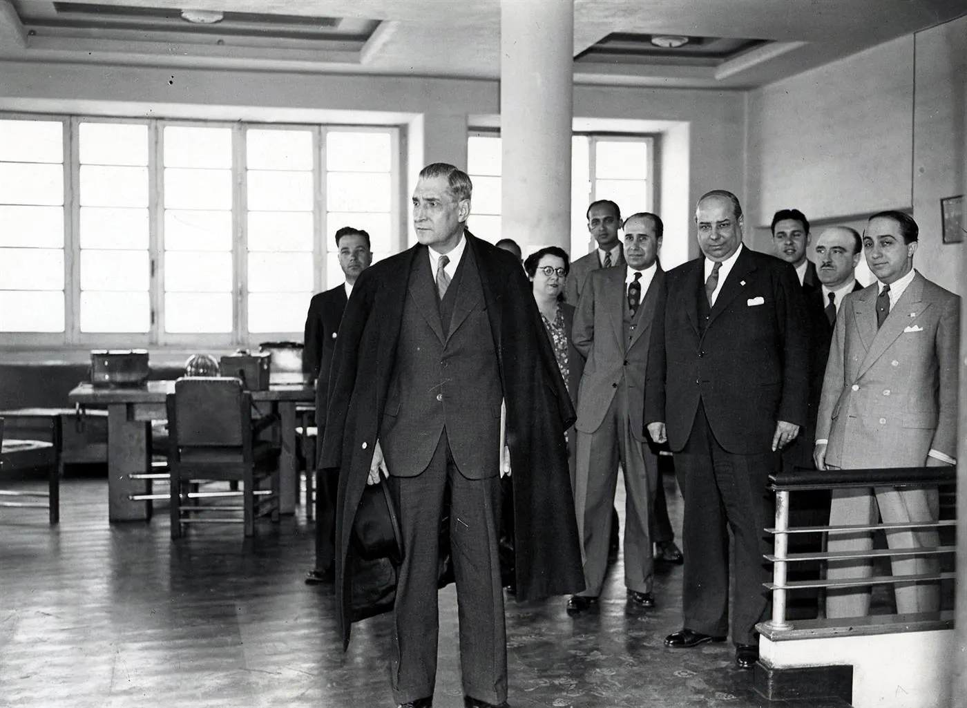 Black-and-white photograph of a tall man in a suit, a dark overcoat draped over his shoulders and a hat in his hand, standing in a bright office with large windows. A group of men in suits and one woman in glasses stand behind him.