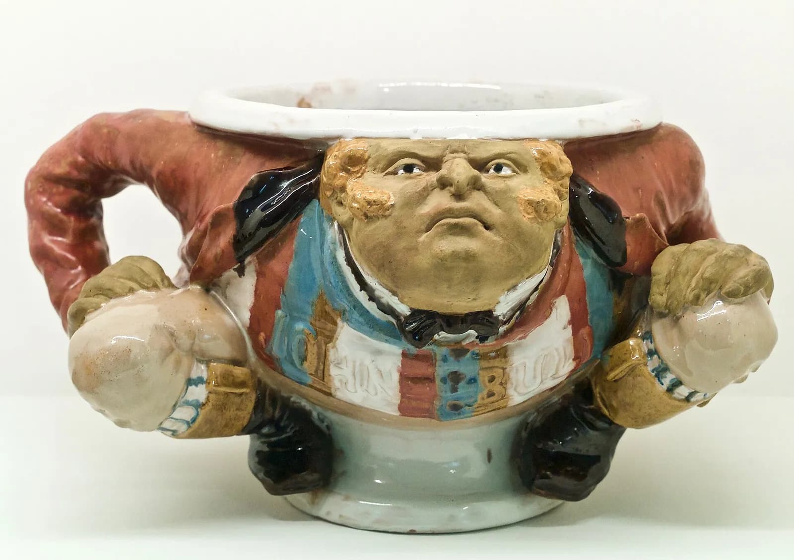 A glazed earthenware chamber pot shaped as a scowling, ginger-whiskered Englishman in a red coat and striped waistcoat lettered JOHN BULL. One arm curves round to form the handle, and his clenched fists jut out at the sides.