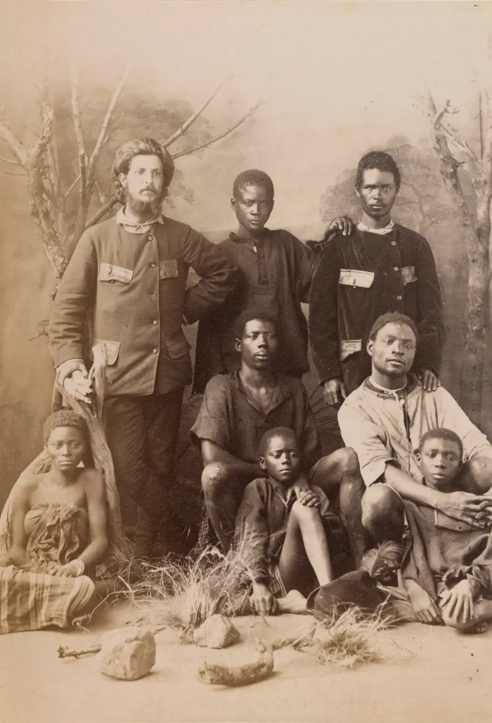 A studio photograph against a painted landscape. A bearded white man in a buttoned jacket stands at the left with one hand on a tree stump; around him stand and sit six young African men and boys and a young woman wrapped in a cloth.
