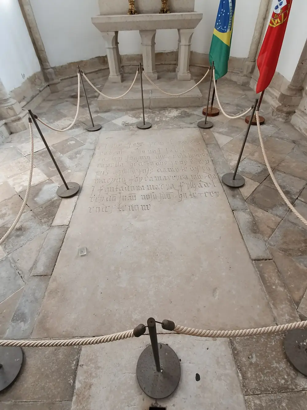 A long stone floor slab carved with a Gothic inscription, roped off with posts in a white chapel. At the far end stand a plain stone altar and the flags of Brazil and Portugal.