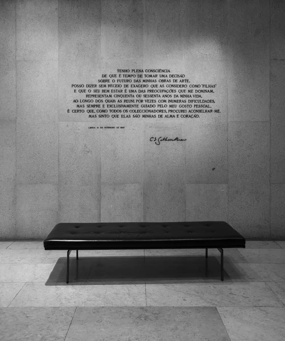 Black-and-white photograph of a pale stone wall carrying ten lines of dark capital letters in Portuguese, a small date line and a handwritten signature reading C S Gulbenkian. A long black leather bench stands on the stone floor in front of it.