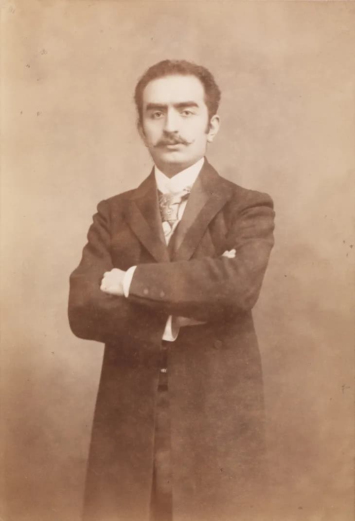 Sepia studio photograph of a young man with dark wavy hair and a thick moustache, standing with his arms folded, in a long dark frock coat, waistcoat and patterned tie.