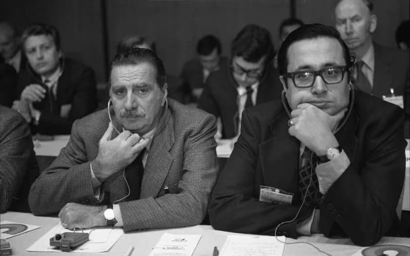 Black and white photograph of two men in suits wearing interpretation earphones at a conference table, both resting their chins on their hands; the man on the right wears thick-rimmed glasses.