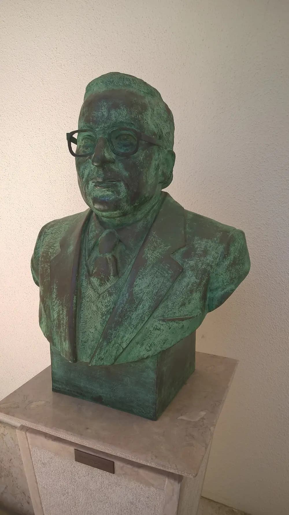 A bronze bust with a green patina of a man in glasses, jacket and tie, on a stone plinth against a white wall.