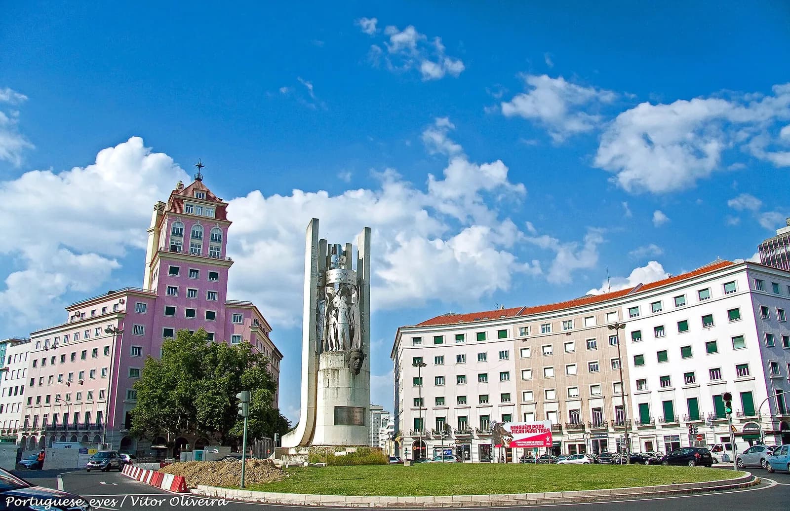 A tall white stone monument with carved figures and a sculpted head stands on a grassy roundabout between pink and cream apartment blocks under a blue sky.