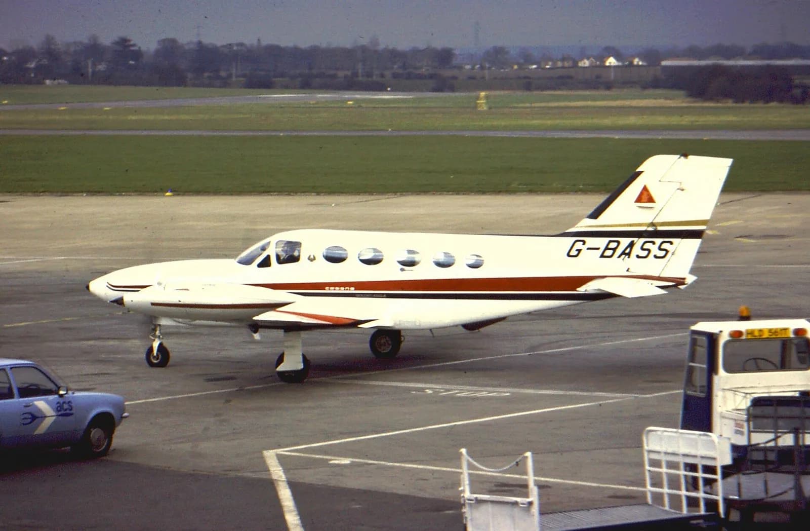 A white twin-engined light aircraft with red and black stripes, a pointed nose and small round cabin windows taxis on an airport apron.