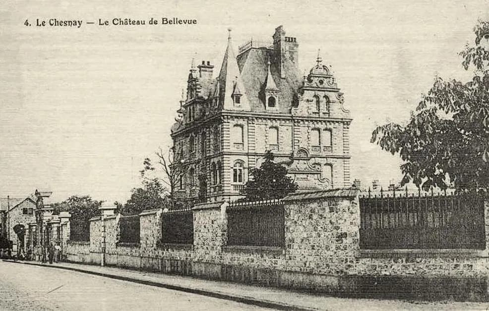 An old postcard of a tall stone mansion with steep slate roofs, dormers and turrets, behind a long stone wall with iron railings on a quiet street. The caption reads 4. Le Chesnay, Le Château de Bellevue.