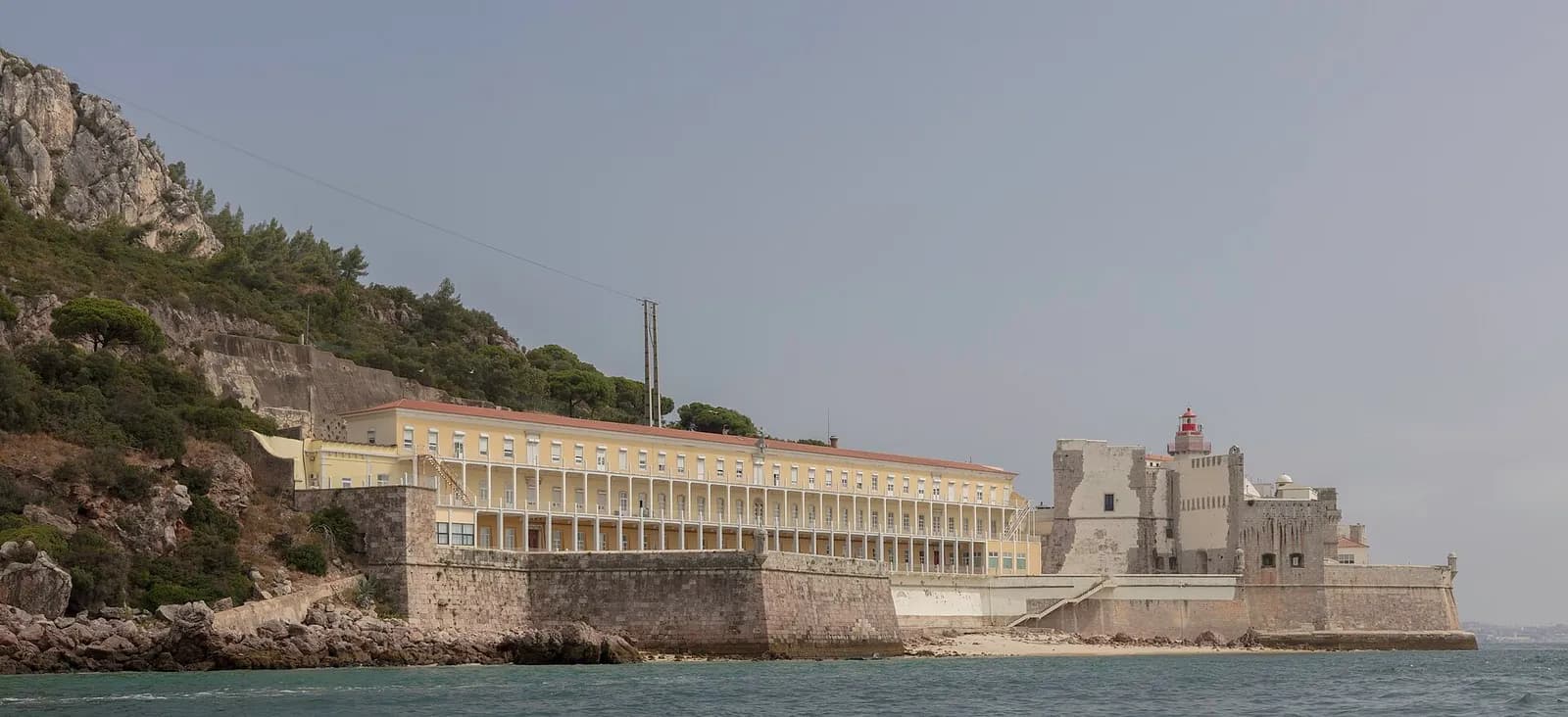 A stone fortress with a red-topped lighthouse stands at the water's edge below a wooded rocky hill. Beside it, behind a sea wall, is a long pale yellow building with three storeys of open galleries.