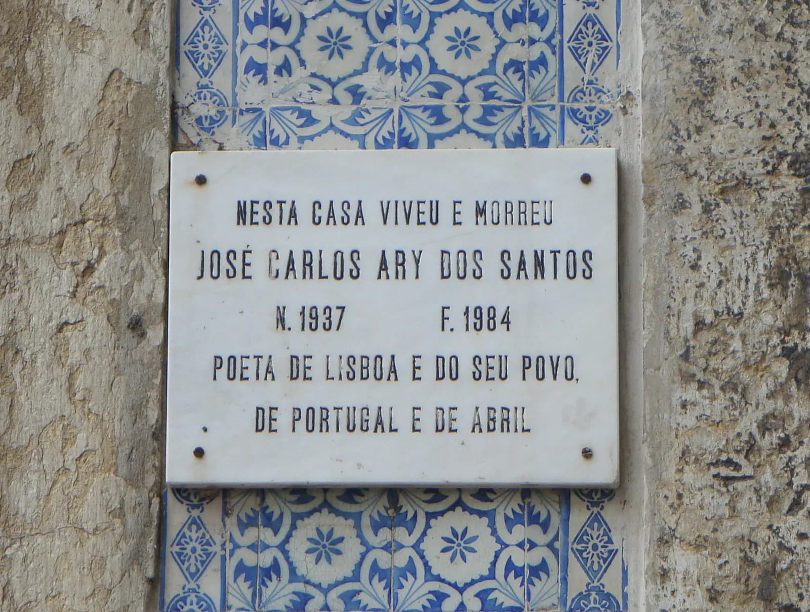A white marble plaque framed by blue and white tiles on a stone wall. Its capital letters read, in Portuguese: In this house lived and died José Carlos Ary dos Santos, born 1937, died 1984, poet of Lisbon and of its people, of Portugal and of April.