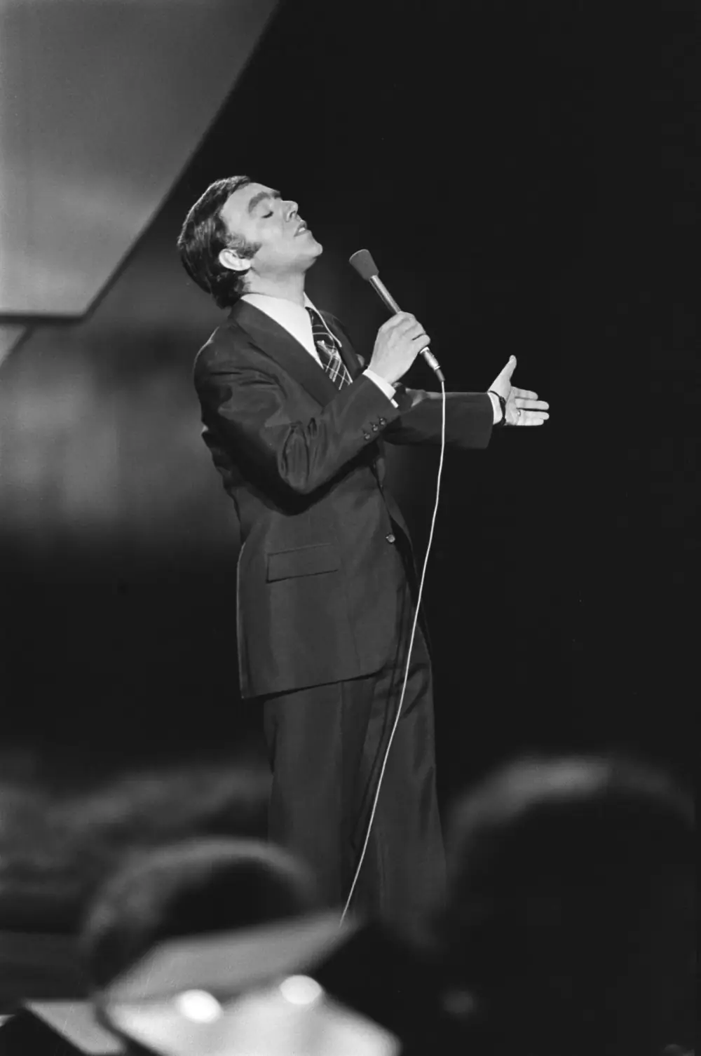 Black-and-white photograph of a dark-haired man in a dark suit and patterned tie singing on a stage, eyes closed and head tilted back, holding a microphone in one hand and stretching out the other. The heads of audience members are blurred in the foreground.
