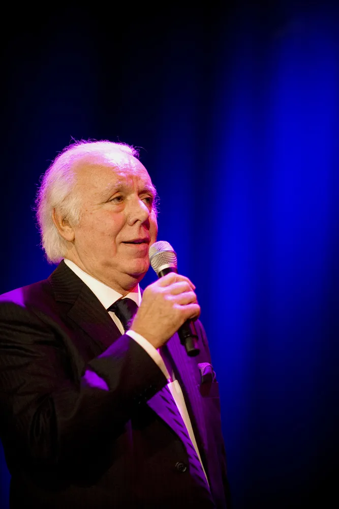 A white-haired man in a dark suit and tie sings into a handheld microphone against a deep blue stage curtain, smiling slightly.