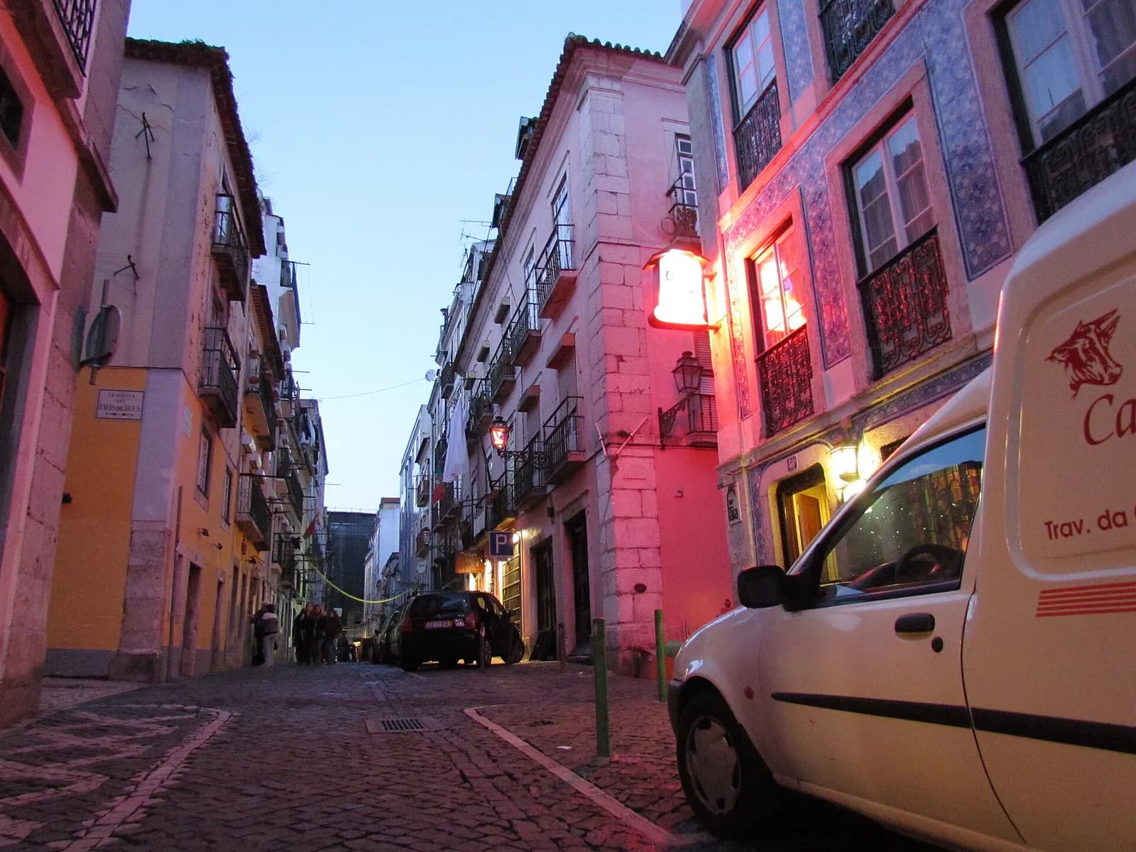 A narrow cobbled street at dusk, lined with tall pastel-coloured houses with iron balconies. A red lantern glows on a tiled façade on the right, and a white delivery van is parked in the foreground.