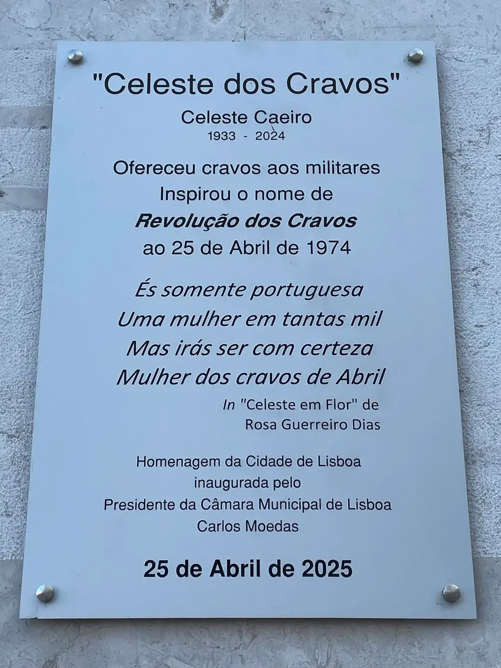A brushed steel plaque on a pale stone wall headed Celeste dos Cravos, with her name, the dates 1933 to 2024, a short text about the carnations and a four-line verse.