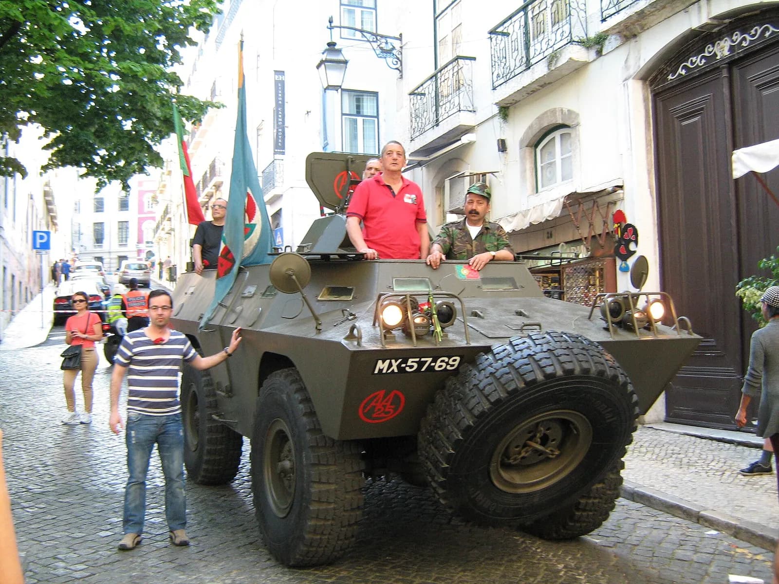 An olive-green four-wheeled armoured car on a cobbled street in front of pale town houses, with a Portuguese flag and a blue regimental flag flying from its rear. Three men stand in its open hull and a man in a striped shirt rests a hand on its side.