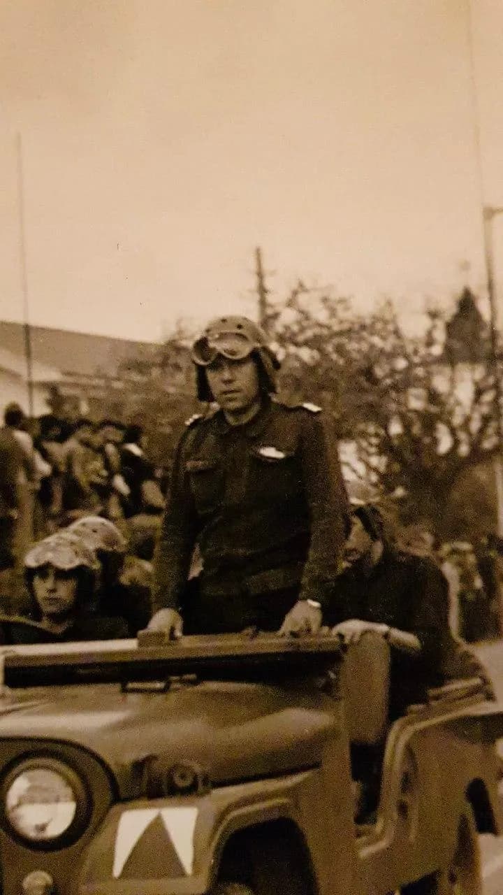 Sepia photograph of a young army officer in a tank crew helmet with goggles, standing in an open military jeep with both hands on the windscreen frame. Soldiers in helmets sit behind him and onlookers stand under trees.
