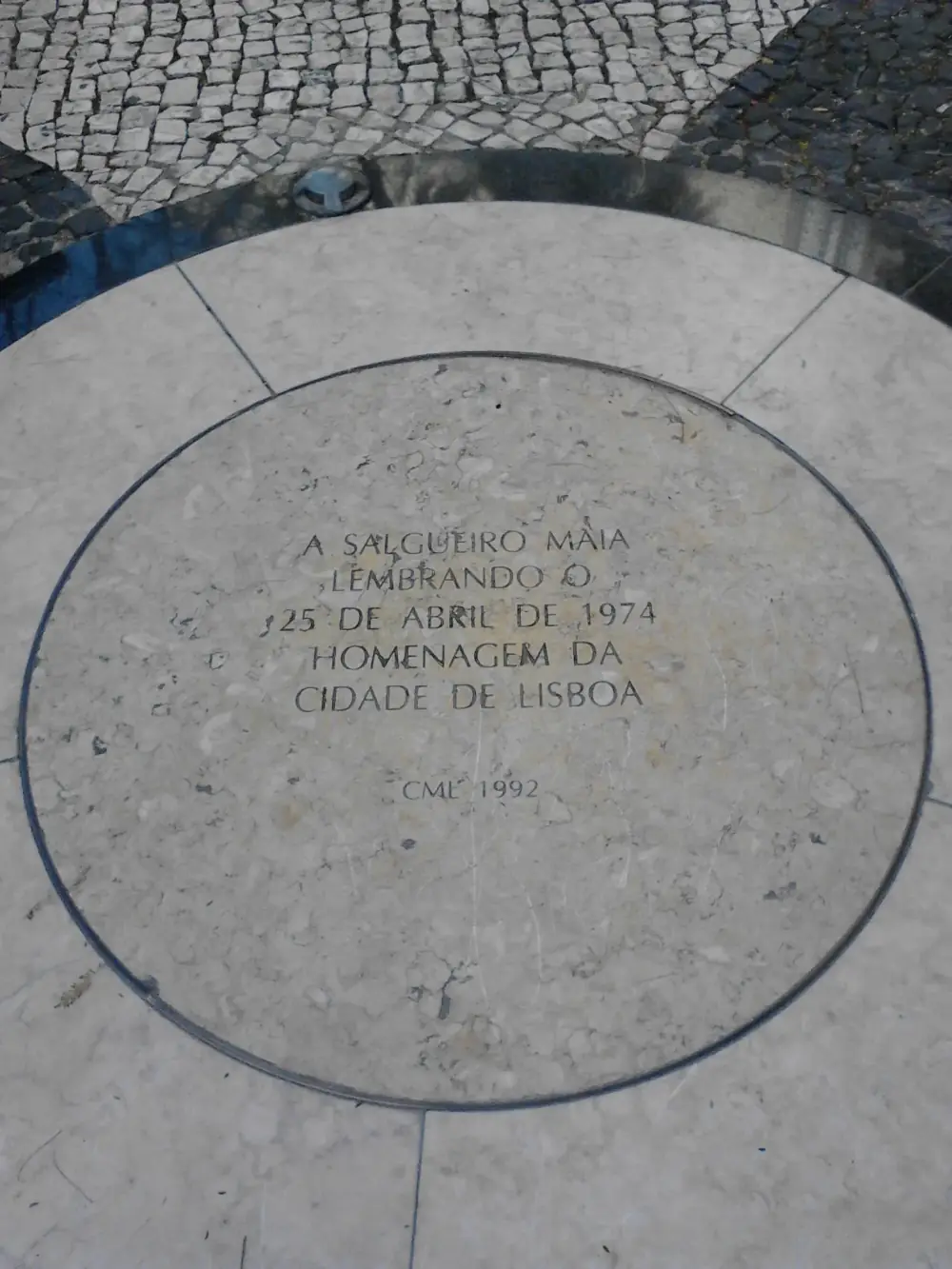 A round slab of pale stone set into a Lisbon pavement of small cobbles, with an inscription in capital letters in its centre and the letters CML and the year 1992 below.