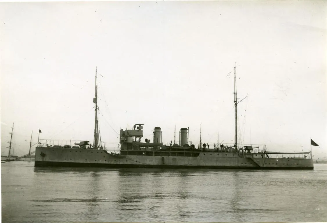 A long grey warship with two funnels and two tall masts lying at anchor on calm water, with sailing ships' masts in the distance.