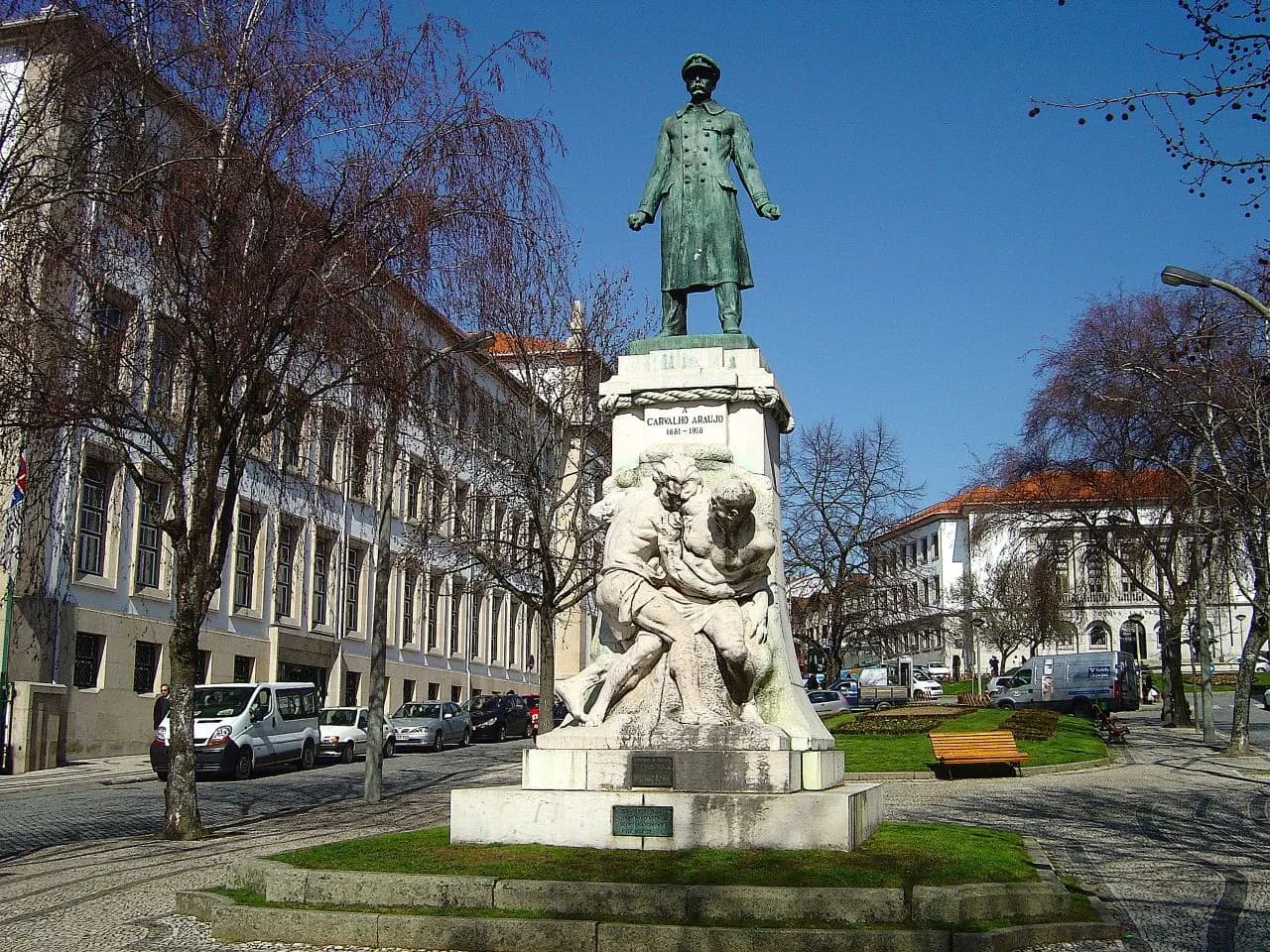 A bronze statue of a naval officer in a long coat, fists clenched, on a tall stone pedestal carved with a struggling sailor, in a garden on a tree-lined avenue.