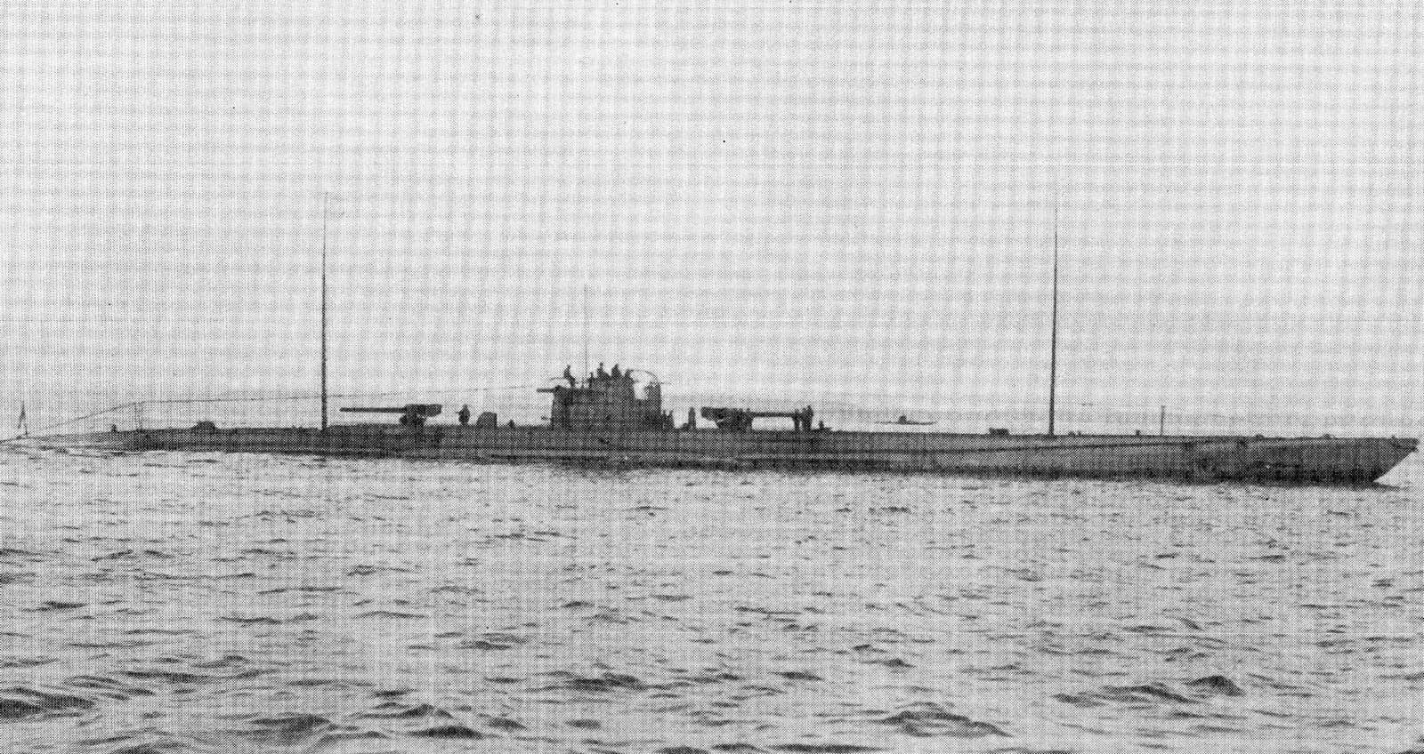 A long, low submarine on a calm grey sea, with a large deck gun forward and another aft of the conning tower, two tall radio masts and a few men on deck.