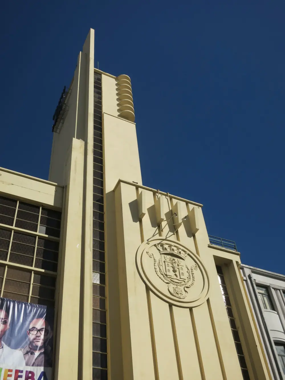 Looking up at a cream-coloured Art Deco tower against a deep blue sky, with a narrow vertical strip of windows, a stack of rounded ledges near the top and a large carved coat of arms in a circle on the wall below.