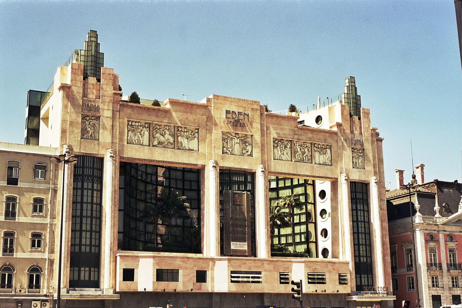 A pale stone Art Deco façade under a clear blue sky, with stepped glass towers at each end, tall dark glazed bays and carved relief panels beneath the lettering Eden Teatro.