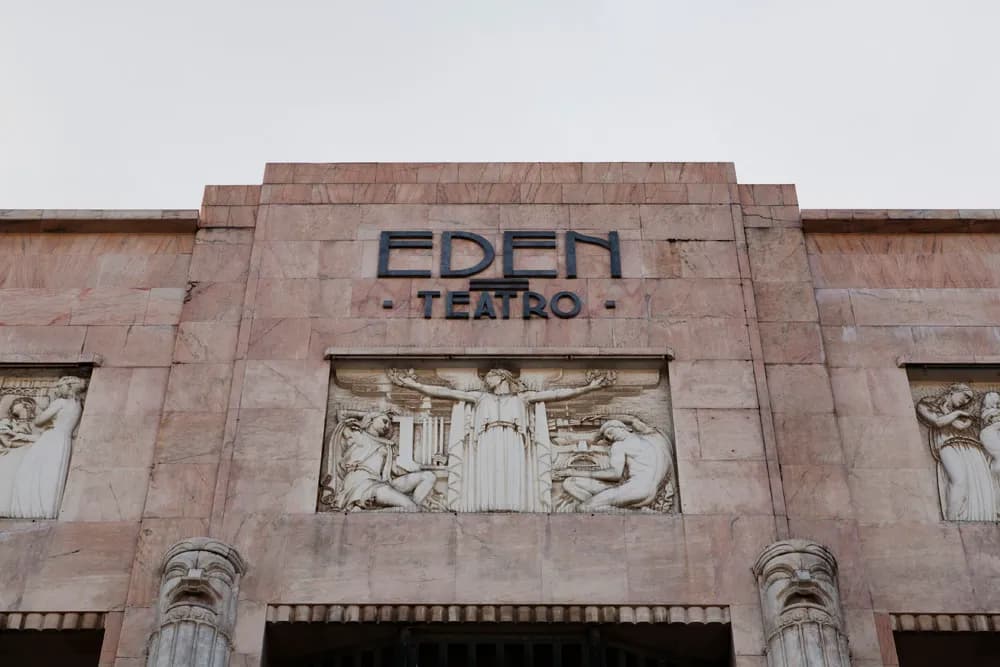 Pink marble façade with the words Eden Teatro in dark metal capitals above a carved stone relief of a robed figure with outstretched arms between two seated figures, with stylised mask-like capitals below.