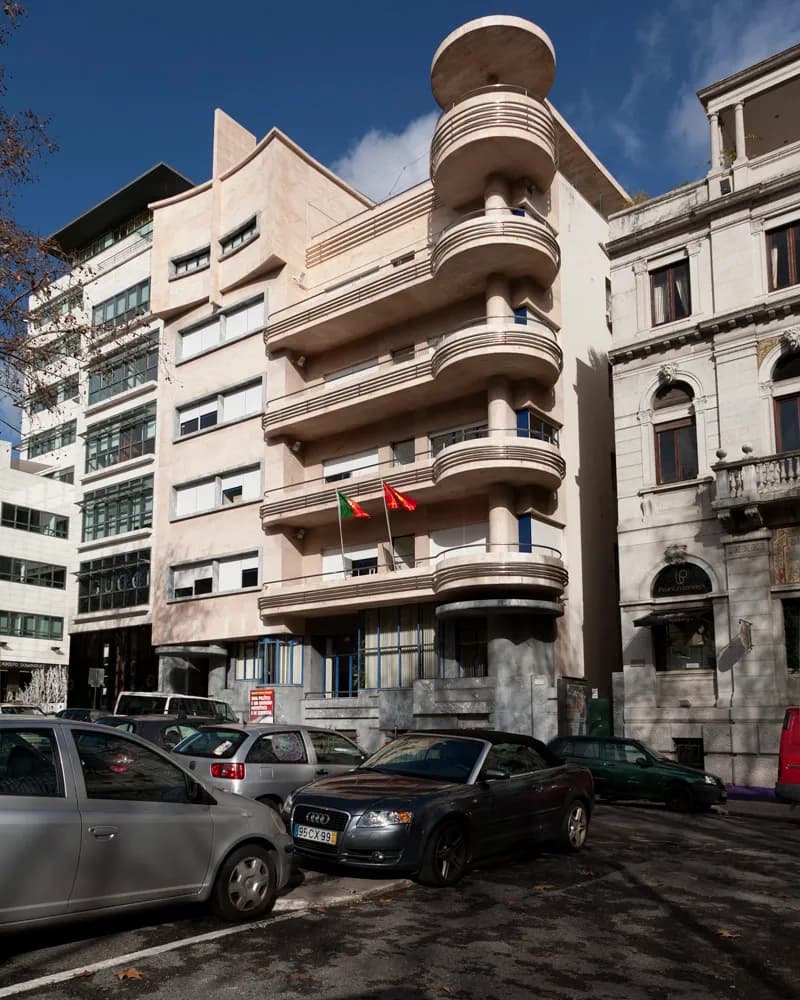 A pale cream modernist building on a city street. On its right, curved balconies are stacked one above another up to a round platform at the top; two flags hang over the entrance and cars are parked in front.