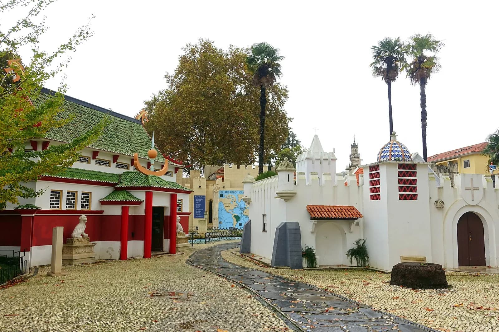 A cobbled path between small buildings: on the left a red and white pavilion with a green tiled Chinese-style roof guarded by stone lions, on the right a white crenellated wall with a small turret topped by a blue and white tiled dome, with palm trees behind.