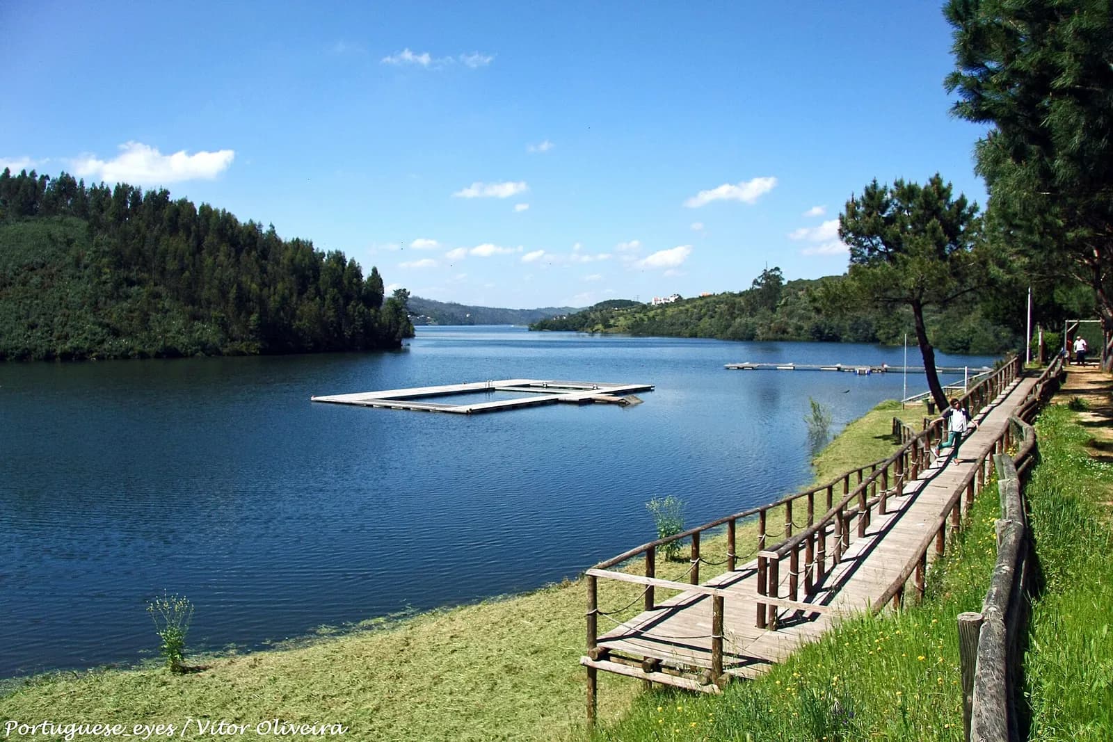 A wooden boardwalk with railings runs along the grassy shore of a calm blue lake, with a child walking on it. A floating swimming platform and a long pontoon sit on the water below wooded hills.