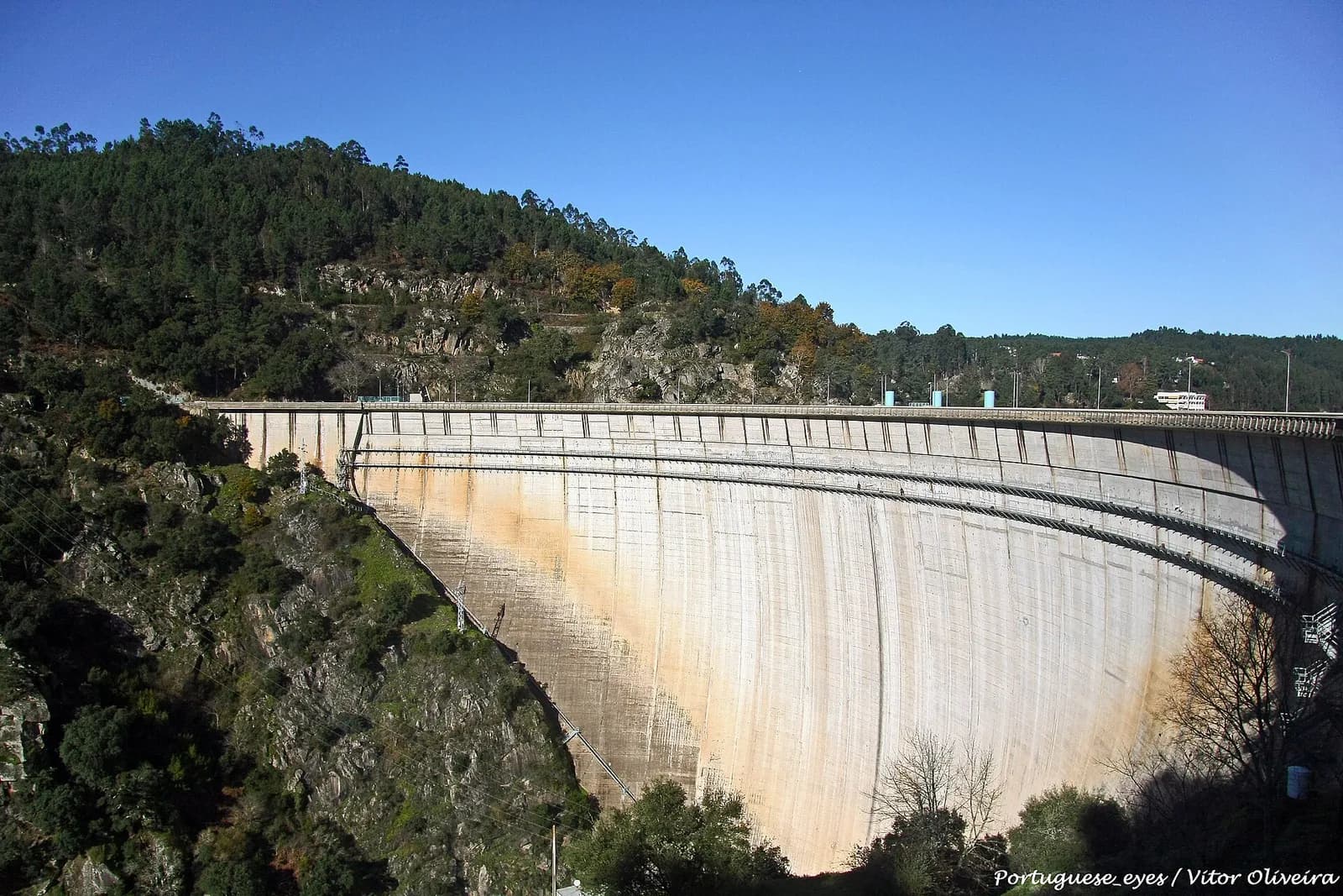 A thin, sharply curved concrete arch dam spans a steep gorge between hills covered in pine and scrub, under a clear blue sky.