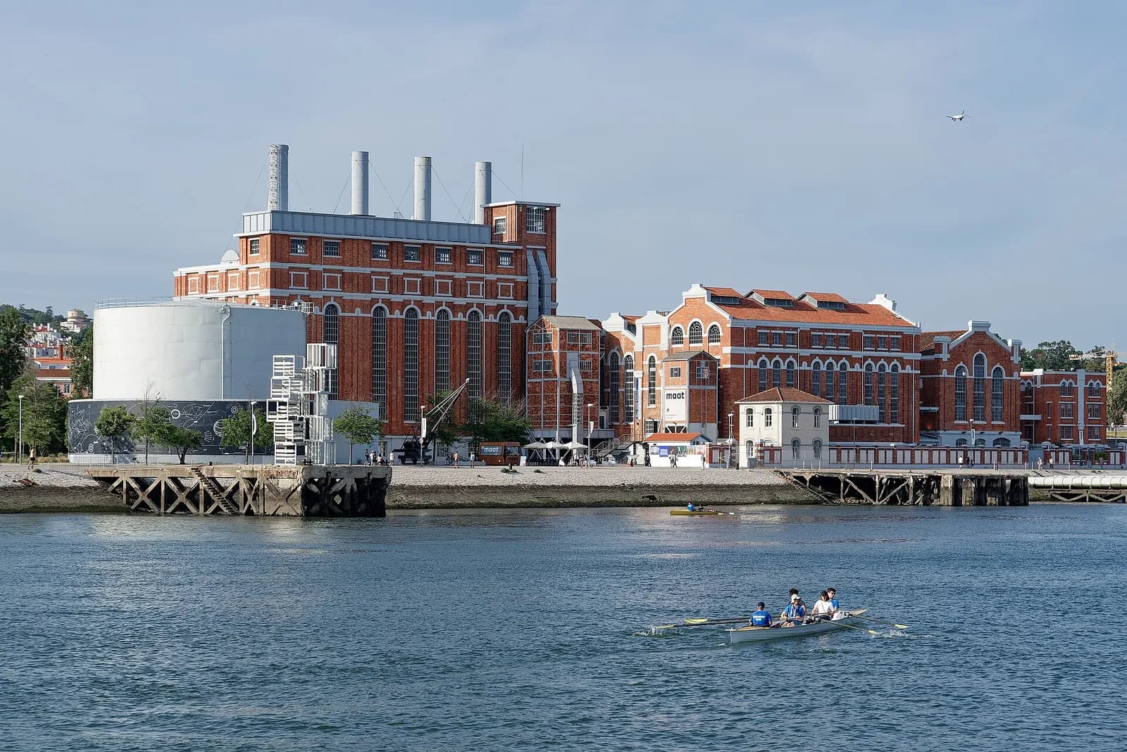 A large red-brick industrial building with four tall chimneys and arched windows stands on the riverfront, next to a white cylindrical tank. A small rowing crew crosses the water in front.