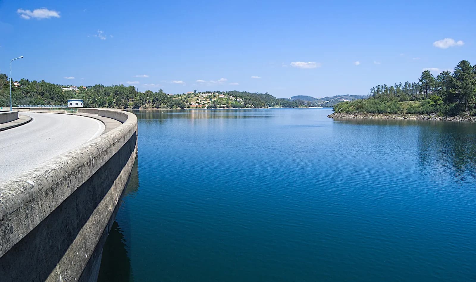 A road curves along the top of a concrete dam behind a low parapet. To the right a wide, still blue lake stretches back between pine-covered hills dotted with houses.