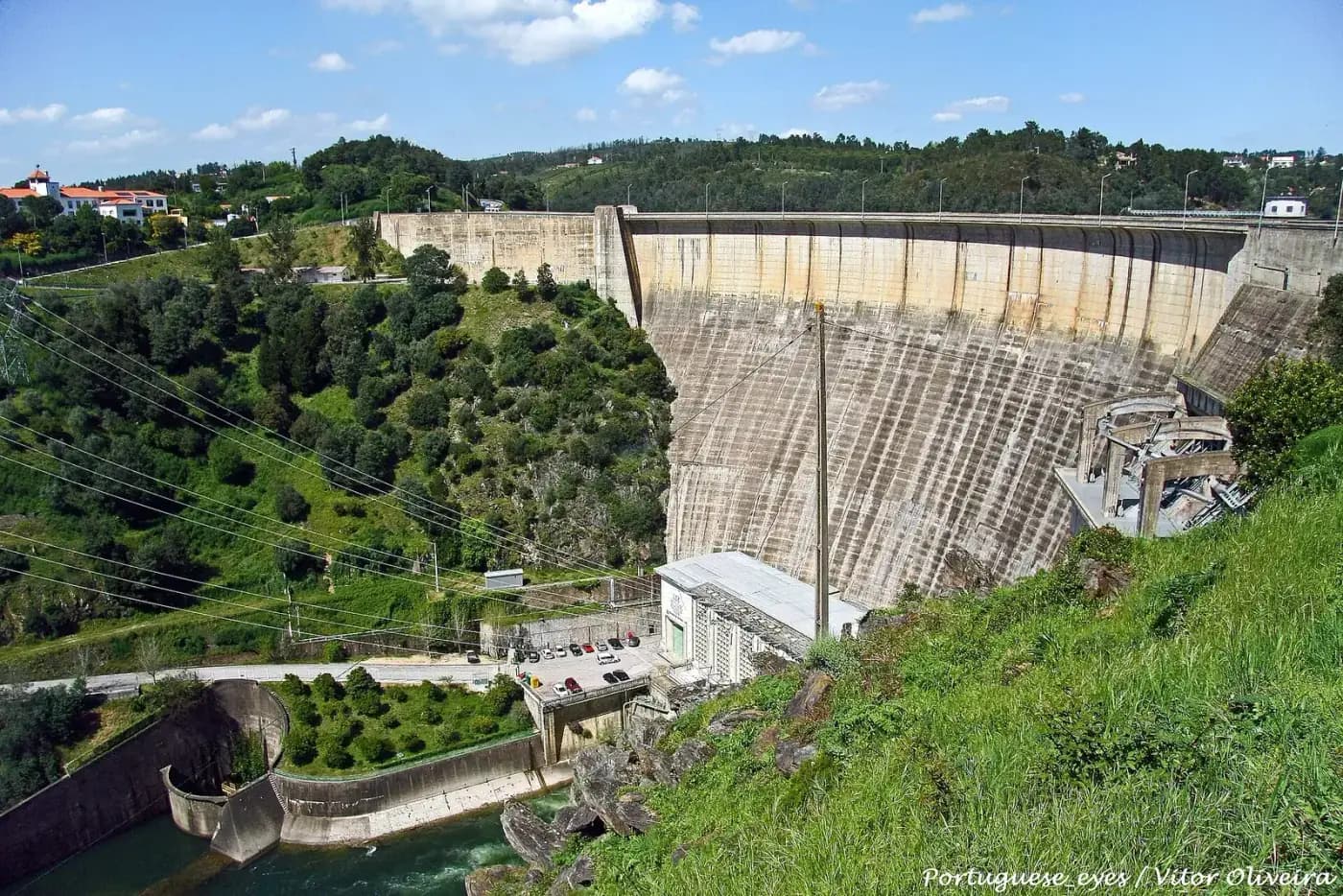 A tall, curved concrete dam wall streaked with water stains closes a wooded valley. At its foot stand a white power-station building, a car park and the outlet channel of the river, with houses on the hilltop to the left.
