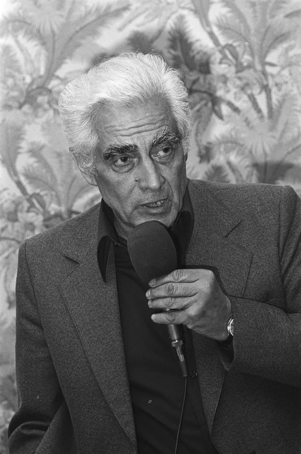 Black-and-white photograph of a grey-haired man in a dark jacket and dark open-necked shirt speaking into a hand-held microphone in front of patterned wallpaper.