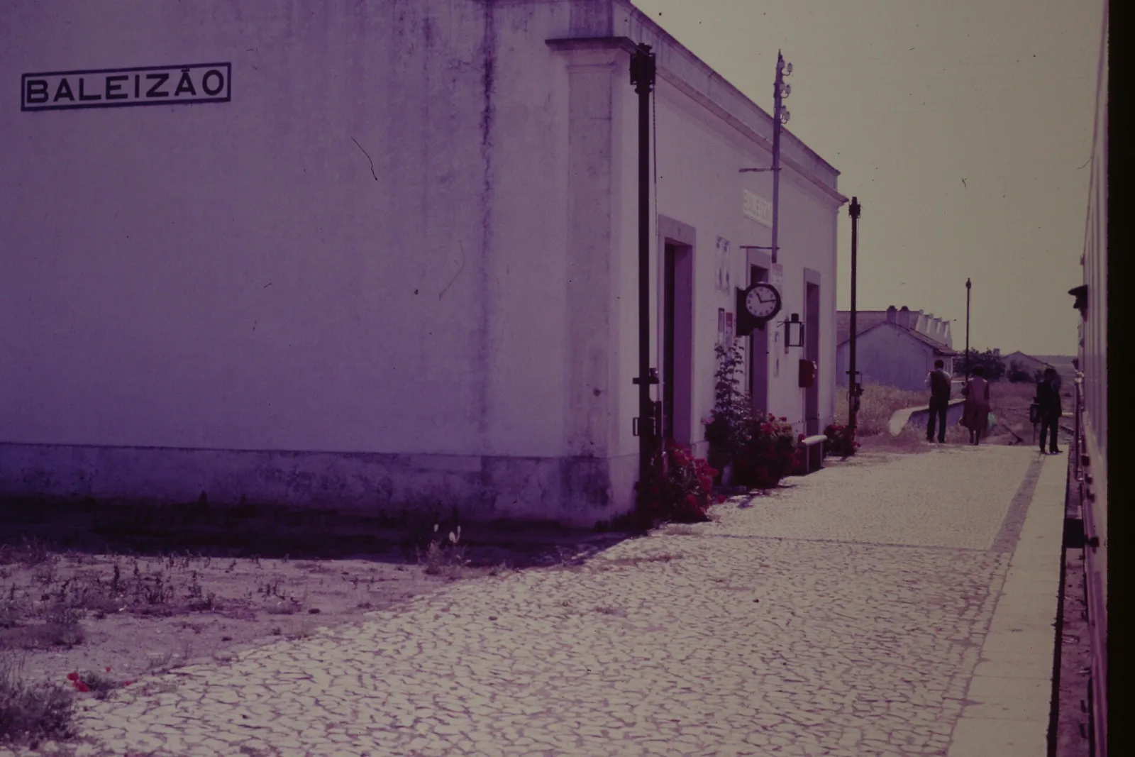 A whitewashed railway station building with a black sign reading Baleizão, a platform clock and red geraniums by the doors, seen from a train standing at a cobbled platform where a few passengers walk away.