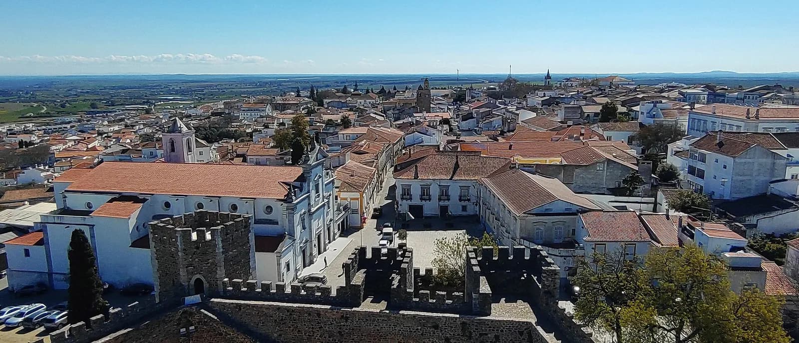 View over the white houses and terracotta roofs of Beja from the castle keep, with battlements in the foreground, a large white church with a baroque front on the left and flat farmland stretching to the horizon.