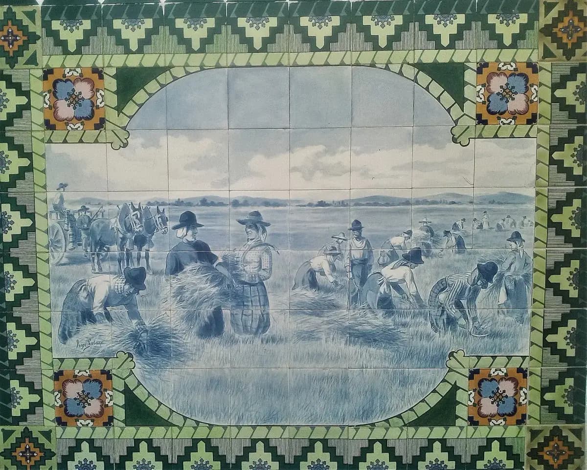 A blue-and-white tile panel in a green patterned border showing a wide flat wheat field. Men and women in broad hats bend to cut the wheat with sickles and gather it into sheaves, while a mule cart waits on the left.