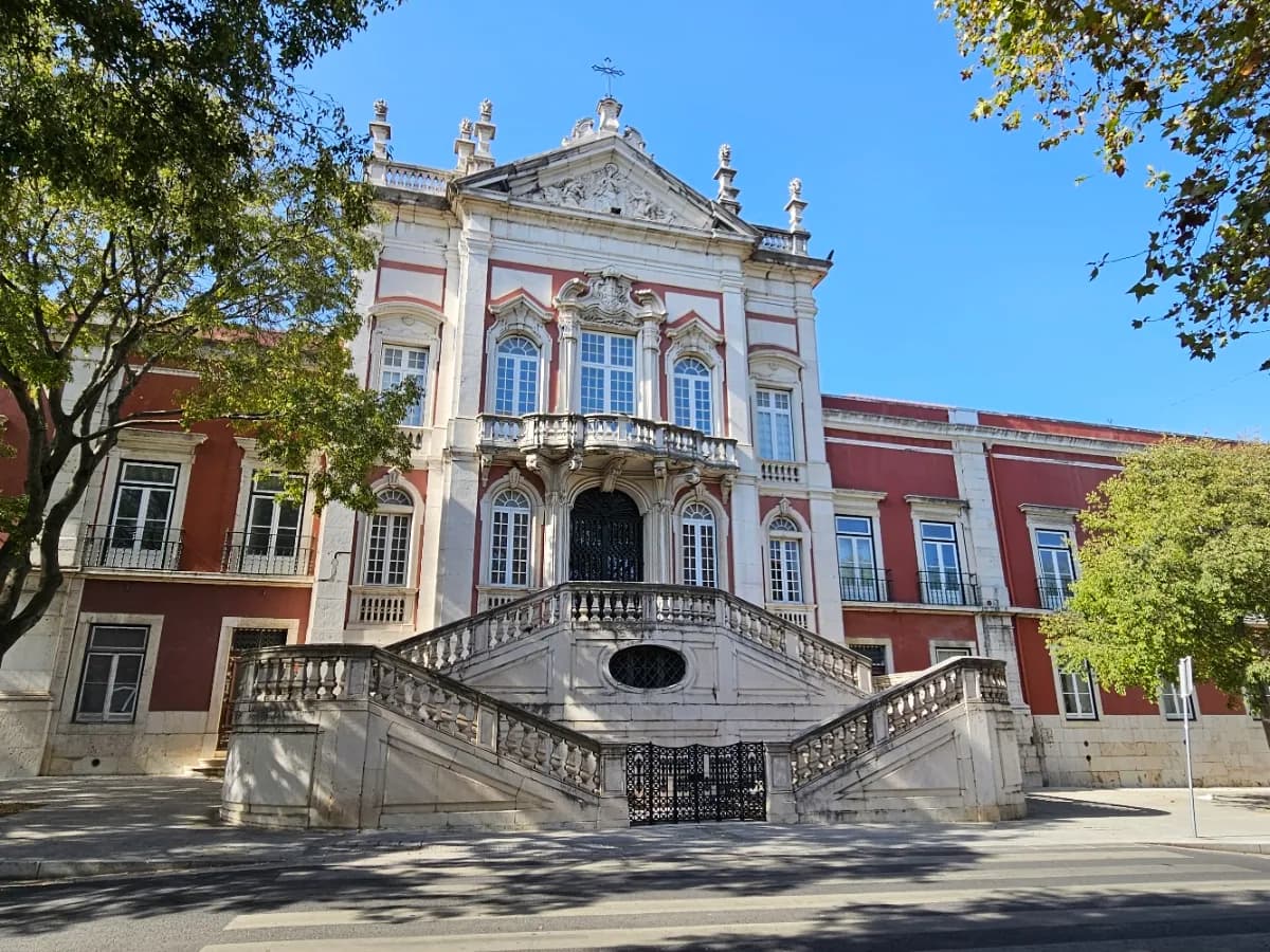 The front of a Baroque palace painted dark red with white stone trim, seen from the street between trees. A double flight of stone stairs with balustrades leads up to a central doorway, and the roofline above has a carved pediment topped by a cross.